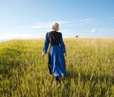 Tours of Lancaster, PA - An Old Order Amish woman in a blue dress and black cape and apron walks in a grassy field on a sunny afternoon